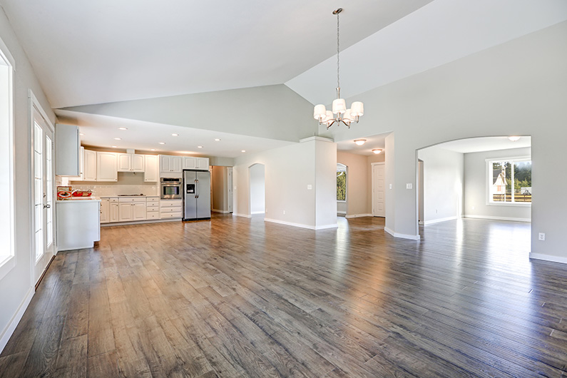 Spacious rambler home interior with vaulted ceiling over glossy laminate floor. Empty light filled dining or living space adjacent to new white kitchen room features pale grey walls. hardwoord flooring, jefferson city missouri